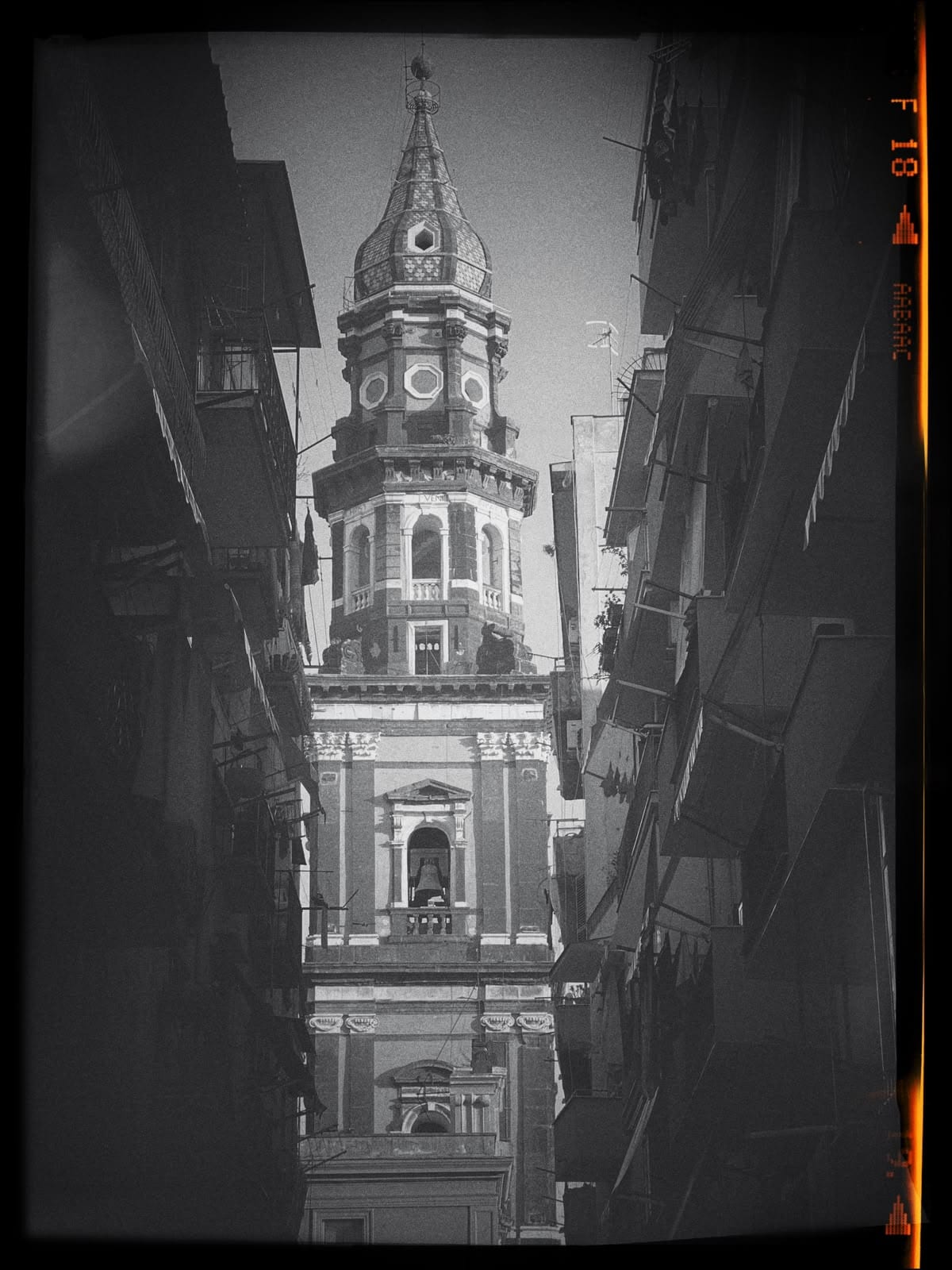 A baroque bell tower seen between buildings, shot in grainy black and white.