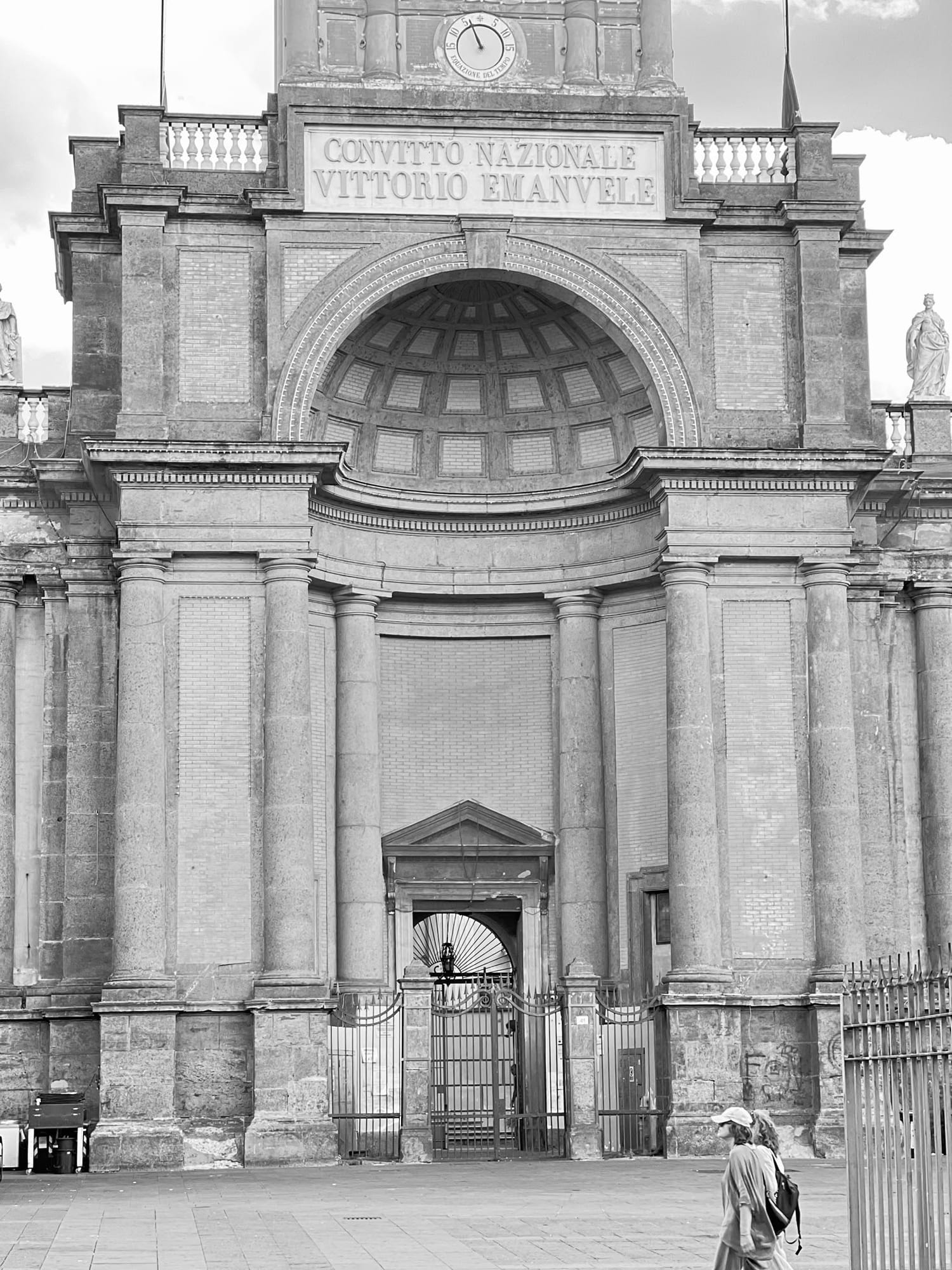 Black-and-white photo of the Convitto Nazionale Vittorio Emanuele facade with a passerby below.