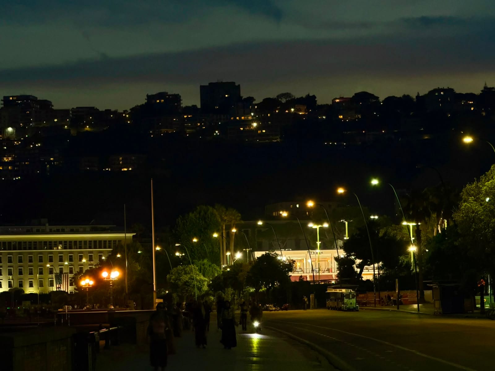 Naples seafront promenade at dusk, streetlights lit, a small tourist train passing.