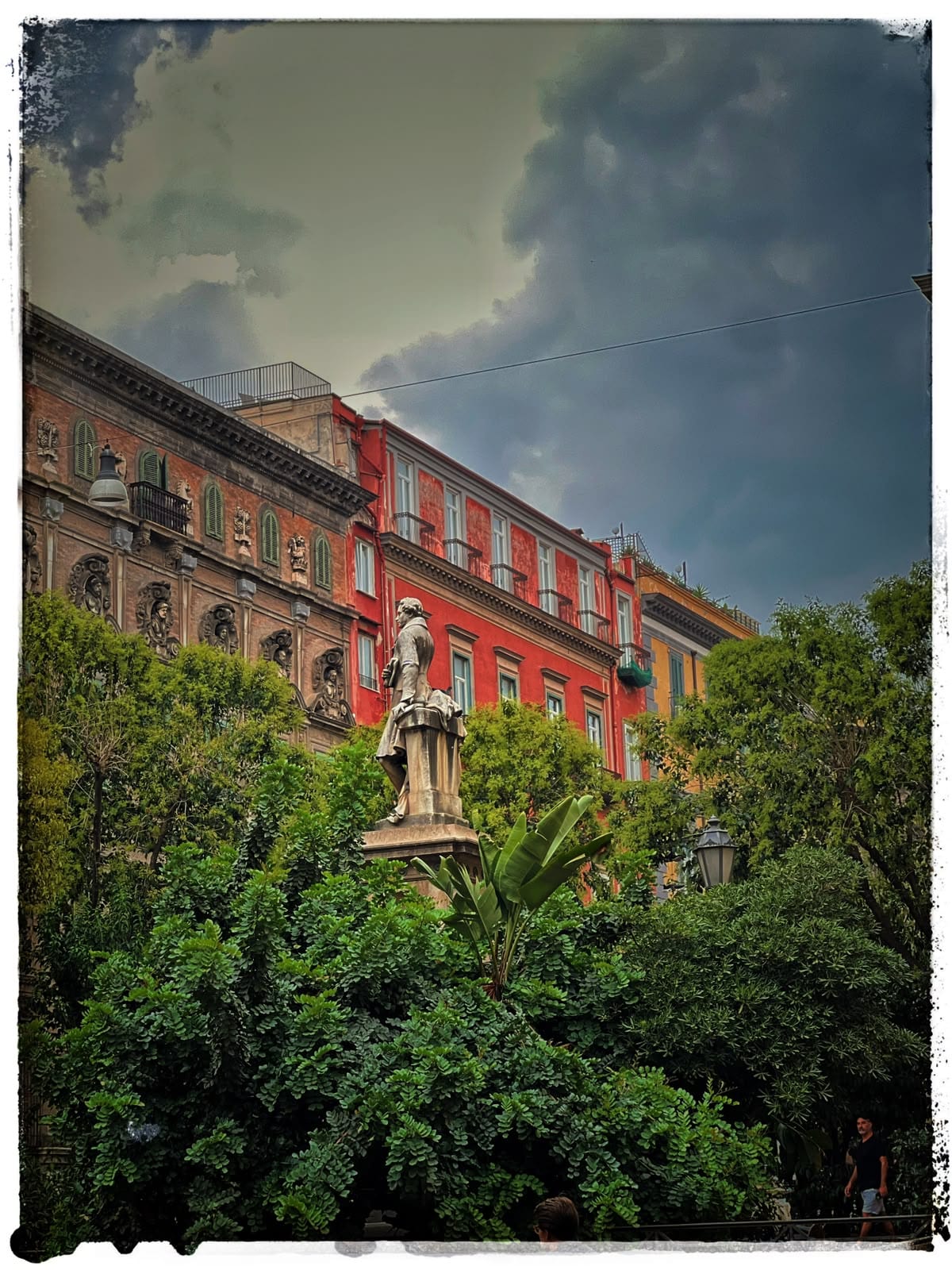 Stone statue set among dense greenery with red and ochre facades behind it.