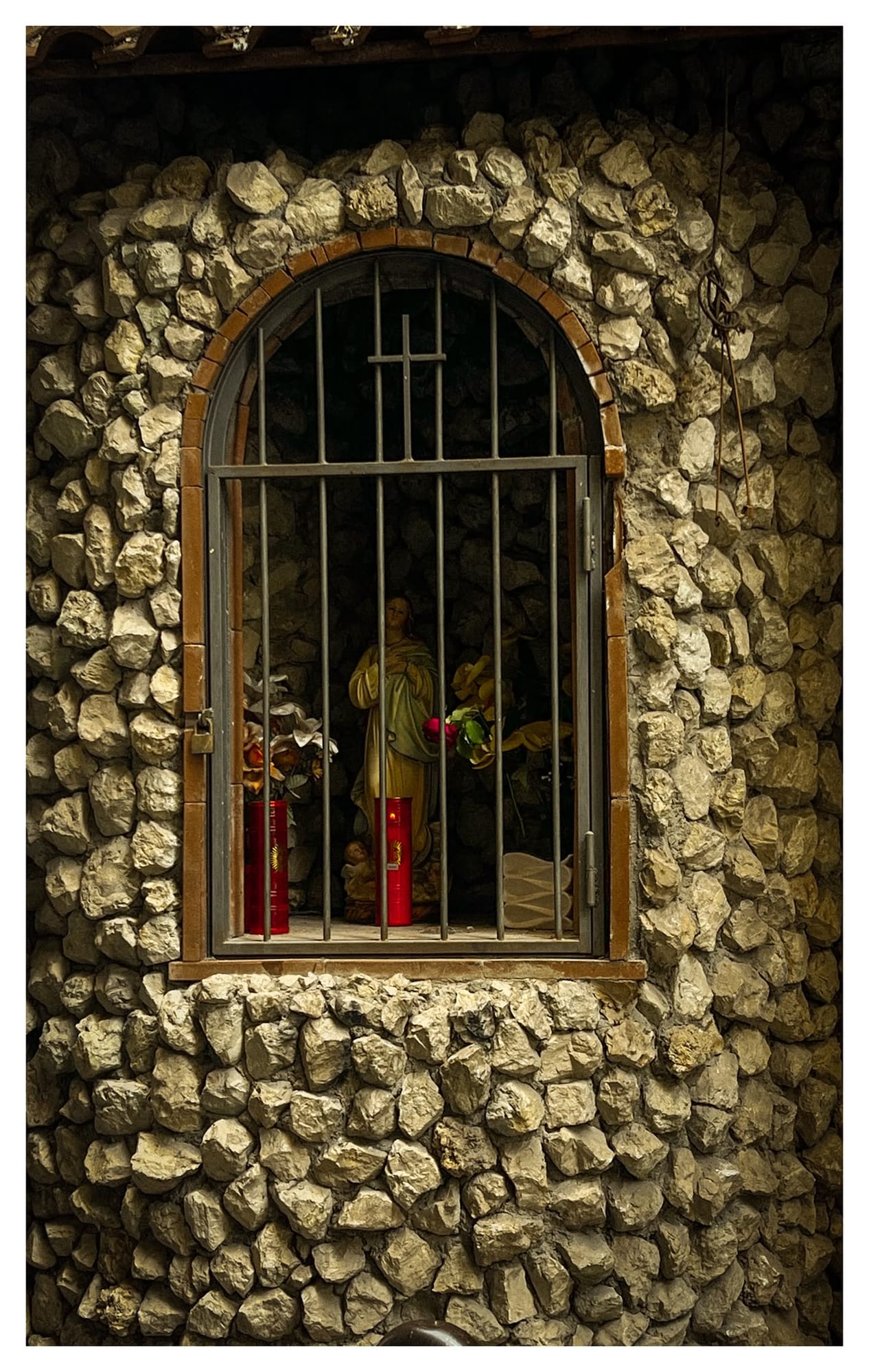 Stone votive niche holding a religious statue, flowers and two lit red candles.