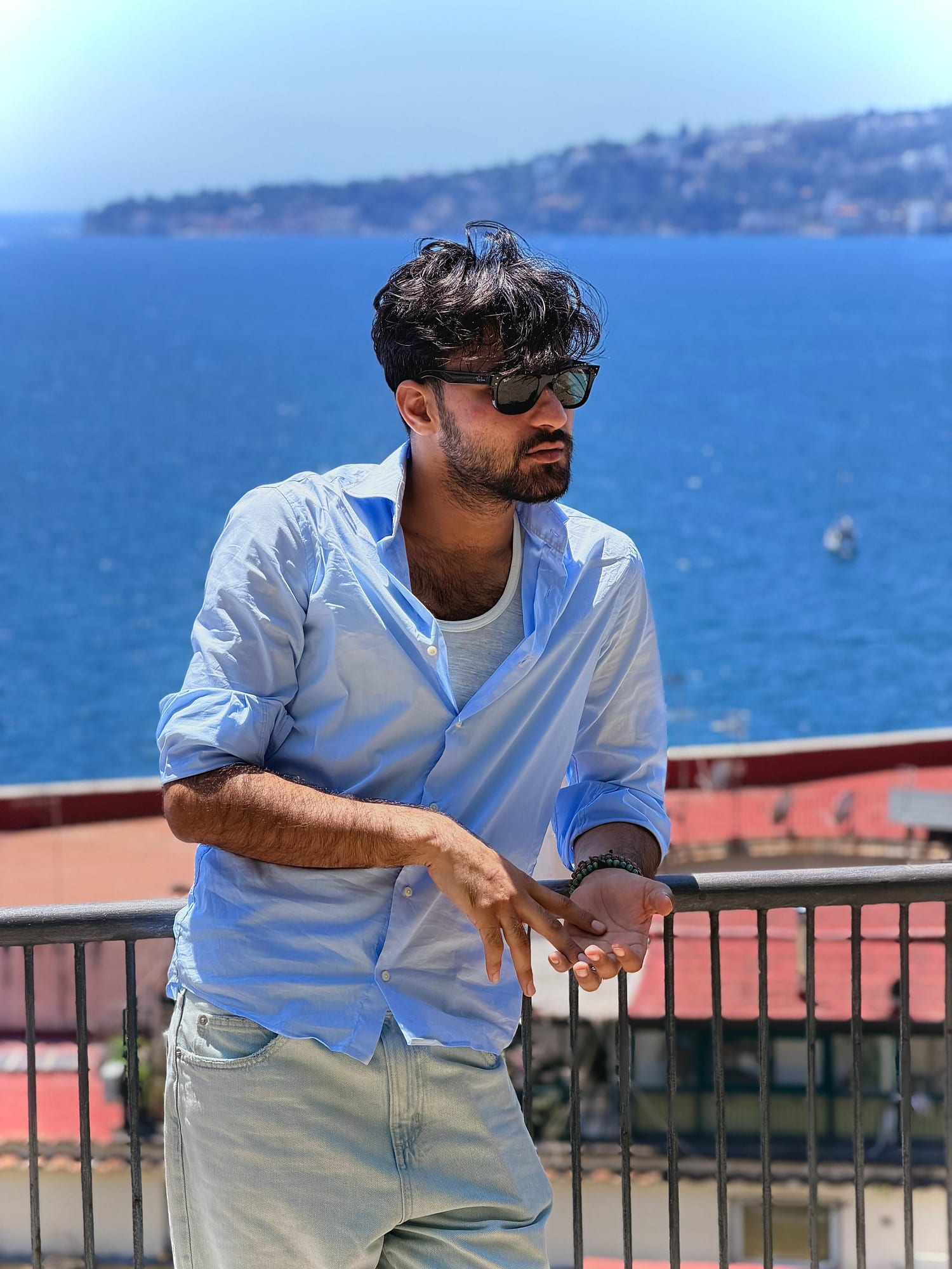 Salar leaning on a railing above the bay of Naples, wearing sunglasses and a light blue shirt, coastline behind him.